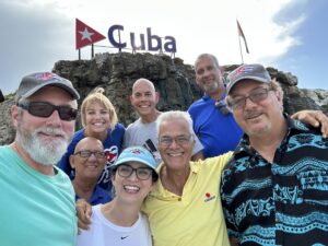 Eight smiling adults pose for a photo in front of a rock with a “Cuba” sign and a red-and-white flag, capturing the spirit of Scenic & Cultivated. | Cubaecology