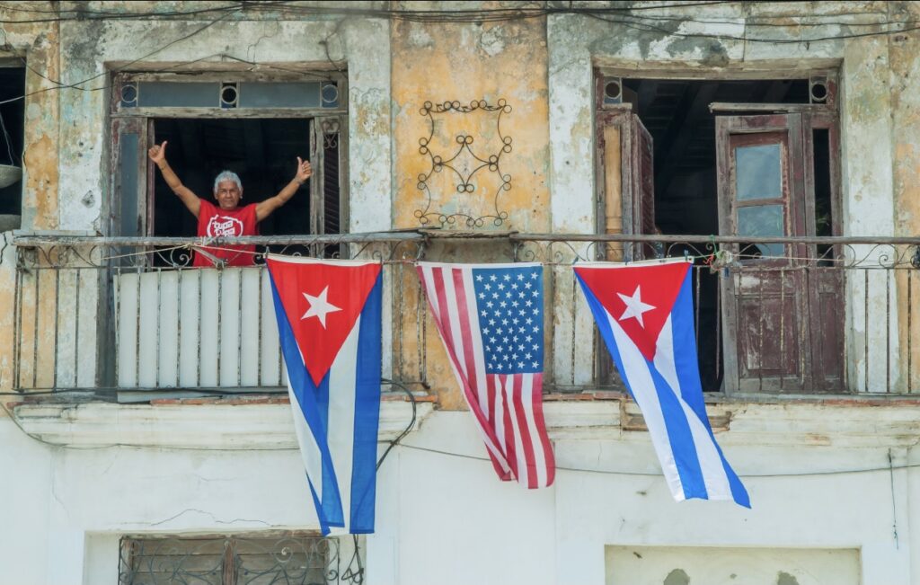 An older person stands on a weathered balcony with two Cuban flags and one U.S. flag hanging over the railing. The building’s paint is peeling. | Cubaecology