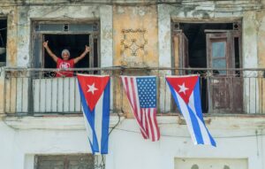 An older person stands on a weathered balcony with two Cuban flags and one U.S. flag hanging over the railing. The building’s paint is peeling. | Cubaecology
