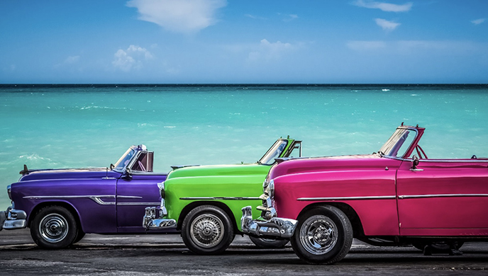 Three vintage convertible cars in pink, green, and purple are parked side by side beside a turquoise ocean under a blue sky. | Cubaecology